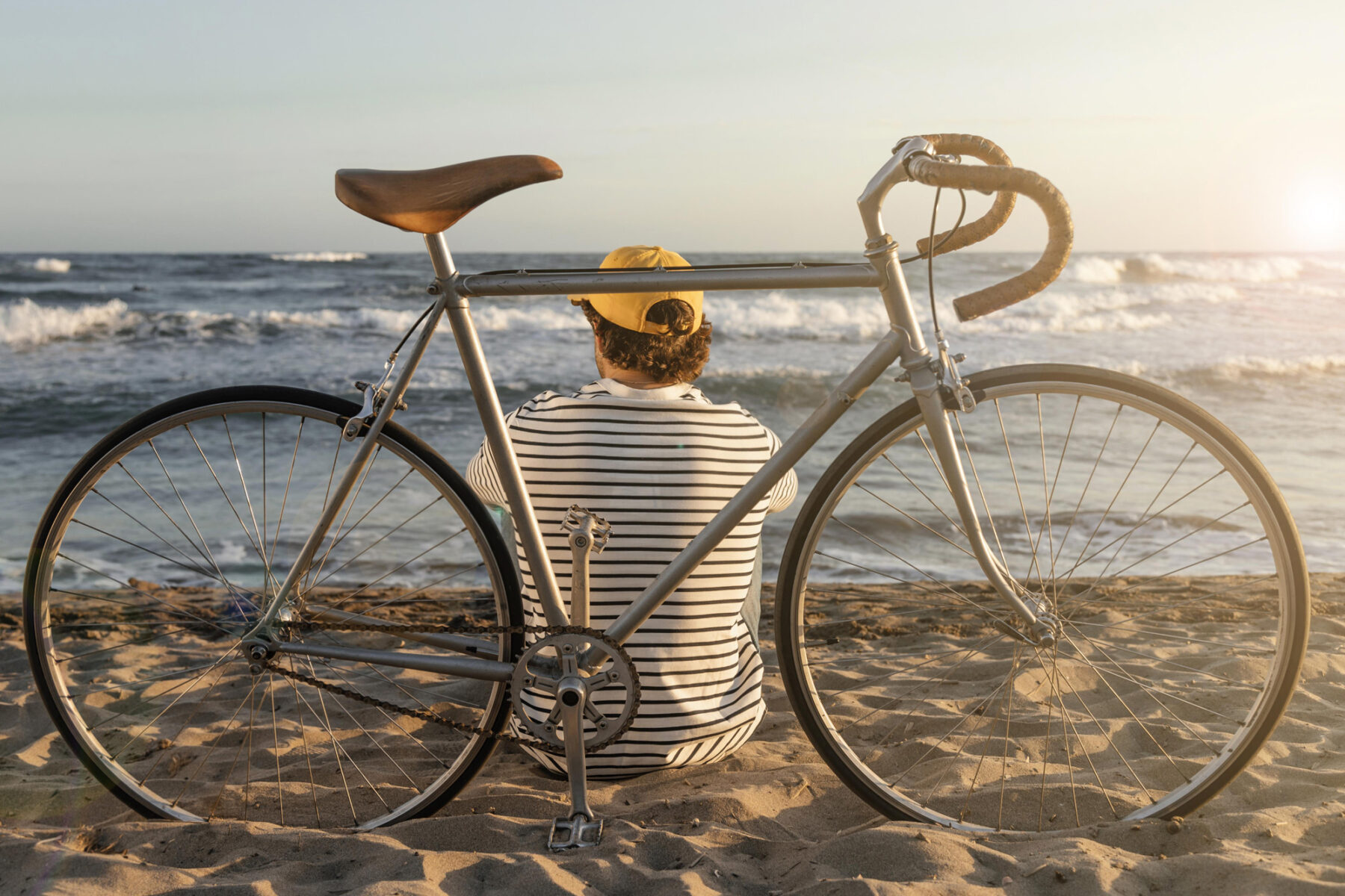 Uomo di schiena seduto sulla sabbia in riva al mare appoggiato alla sua bicicletta