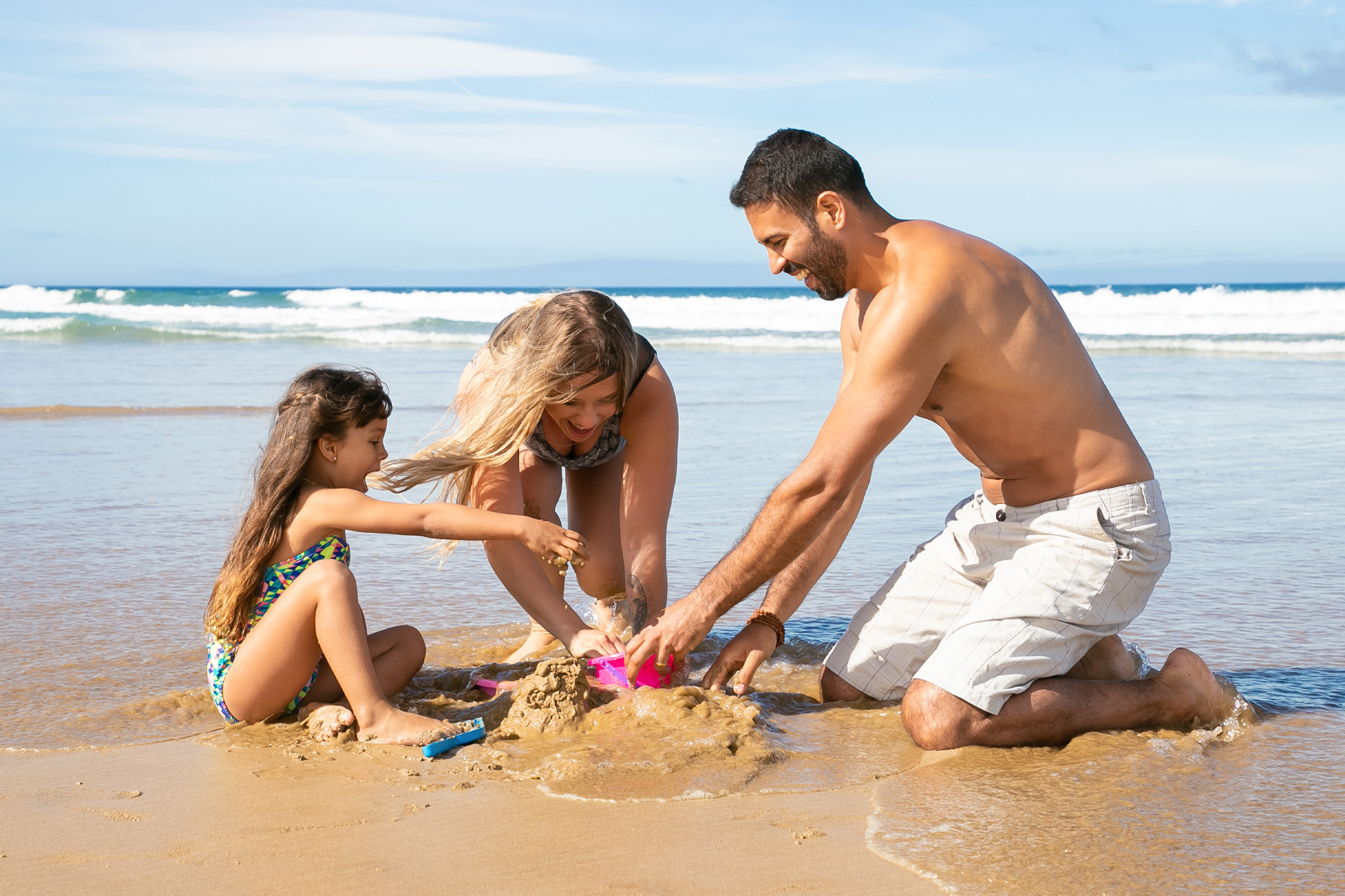 Mamma, papà e bambina in ginocchio sul bagnasciuga che giocano con la sabbia